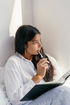 A thoughtful young woman holding a pen and notebook, sitting by a window filled with natural light.